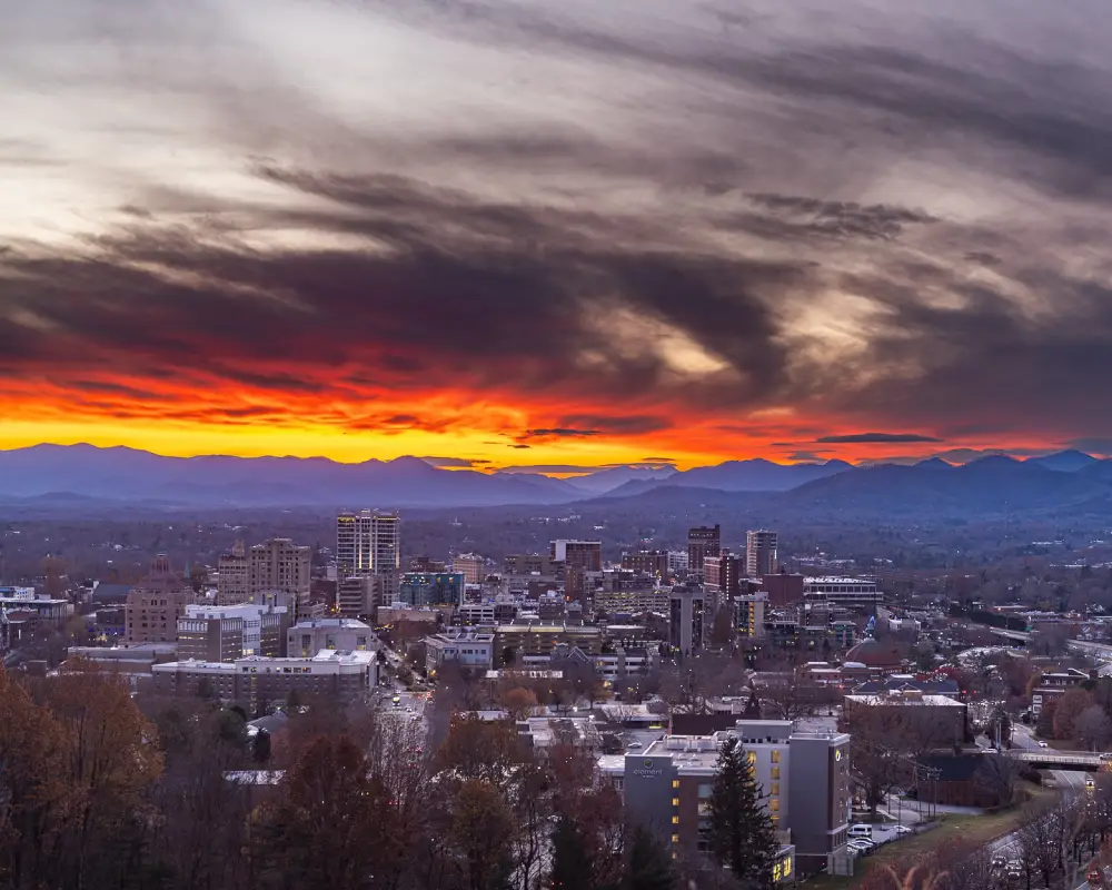 Panoramic view of Downtown Asheville skyline at sunset, with buildings, autumn foliage, and the Blue Ridge Mountains under a fiery orange sky.