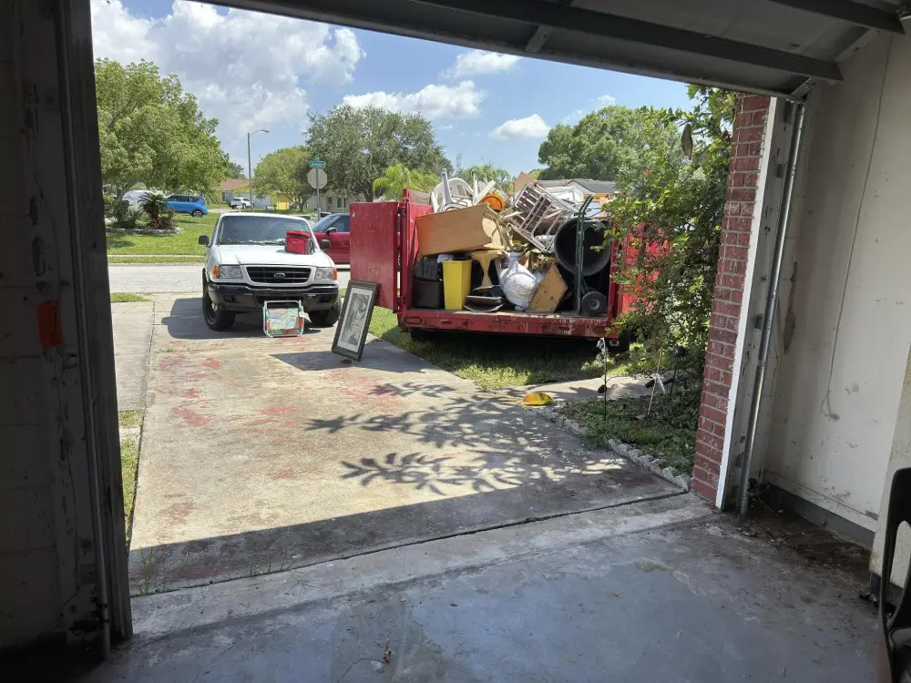 View from open garage of white truck with red dumpster filled with boxes, chairs, wheelbarrow, and debris on a sunny suburban driveway.