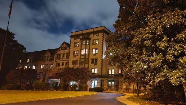Night view of a multi-story historic building with illuminated windows, American flag, curved driveway, green lawn, and surrounding trees under cloudy skies.