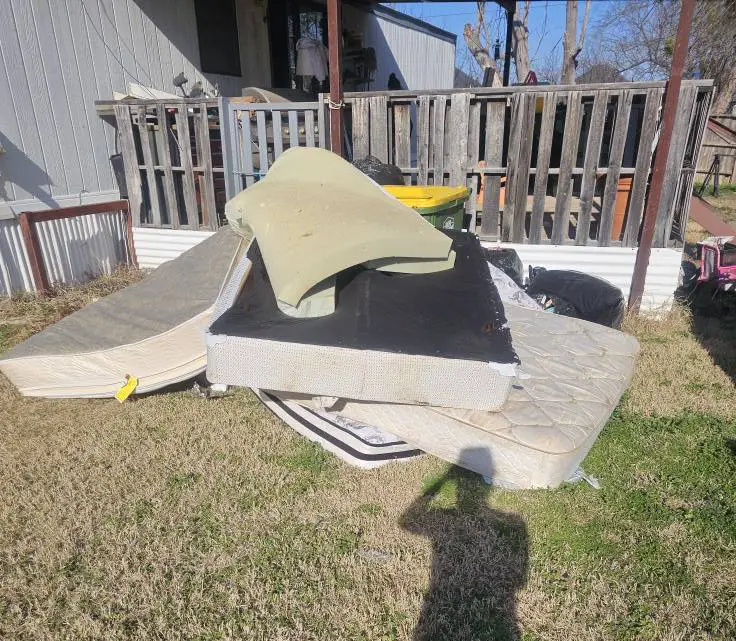 Stack of old mattresses, box springs, and cushions on grass beside a wooden fence and house under clear skies.