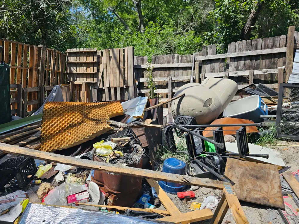 Pile of junk in a fenced yard including barrels, wooden pallets, foam panels, crates, buckets, and scattered trash under trees on a sunny day.