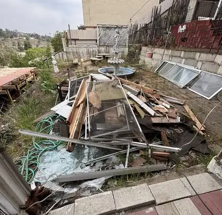Pile of assorted junk in a backyard, including wood scraps, broken glass windows, metal frames, and a green hose, on a concrete patio with overgrown grass.