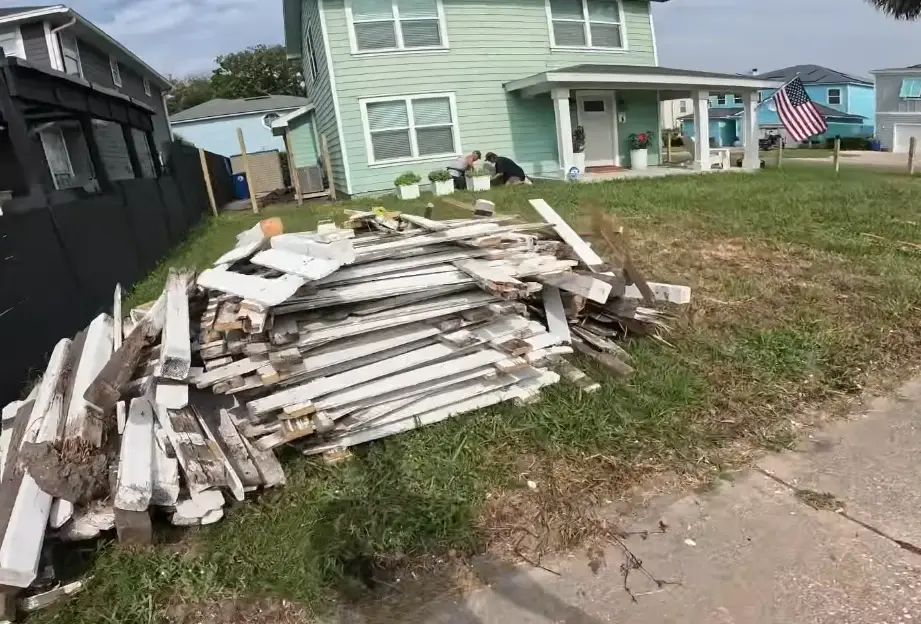 Two junk removal crew members loading a large pile of old fence boards and wood debris outside a residential property in Asheville NC